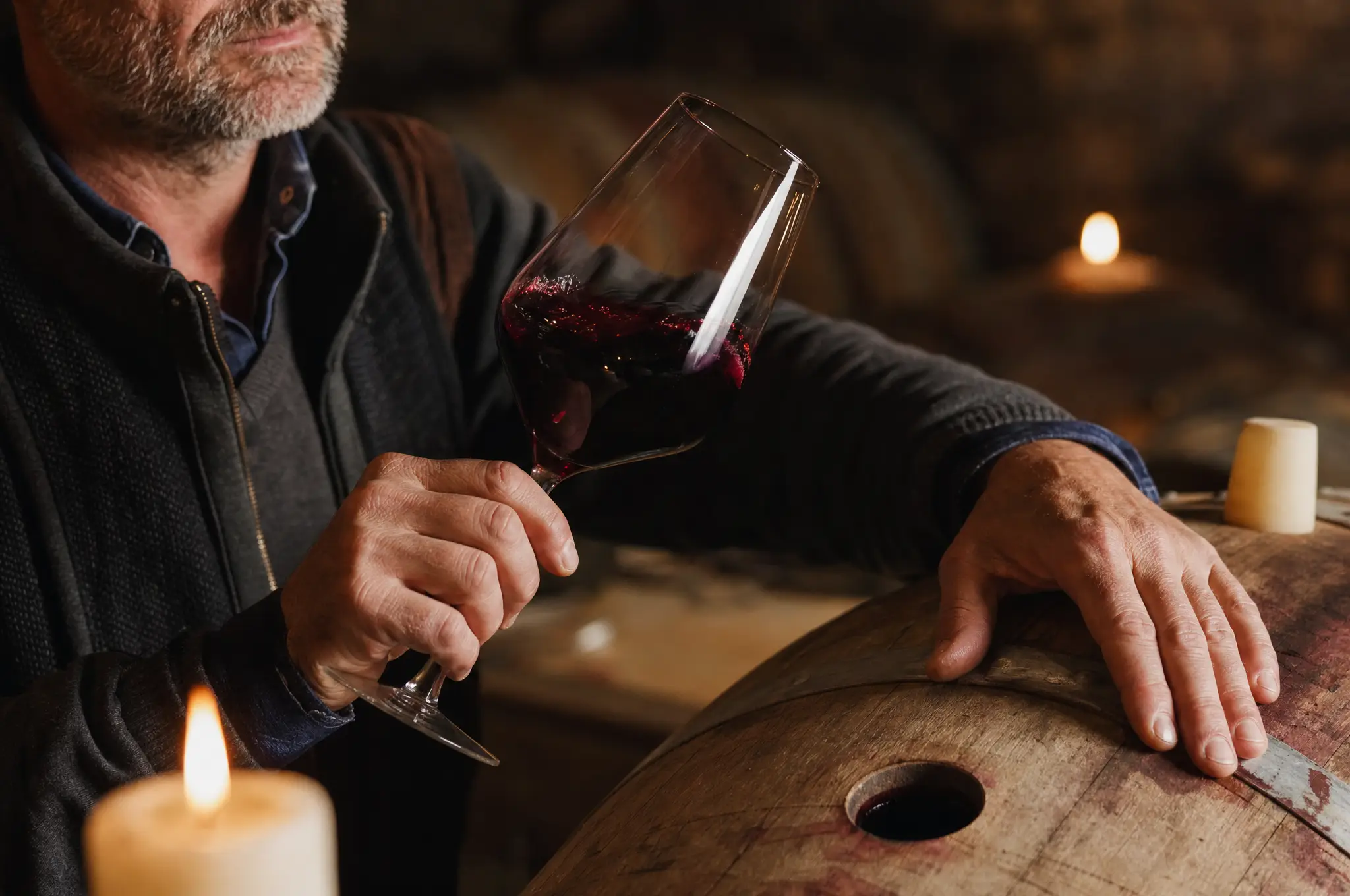A winemaker swirling a glass of red wine beside a barrel in the cellar